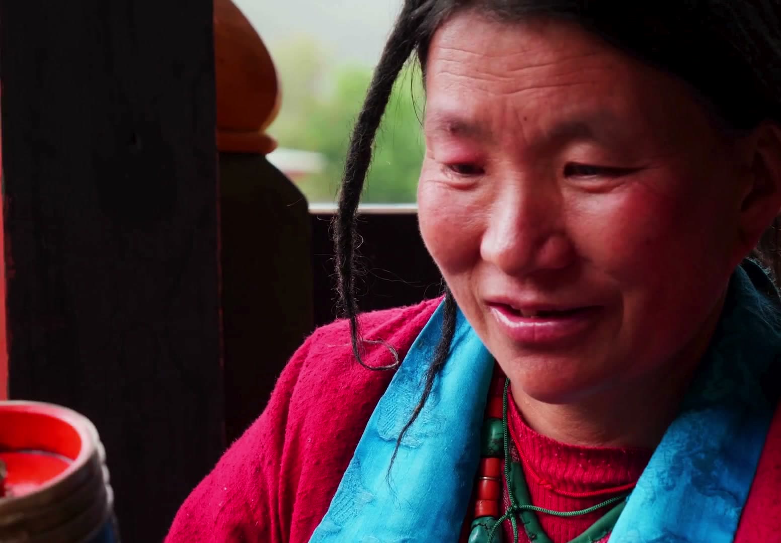 A smiling Bhutanese woman in a red tego and turquoise scarf, at home